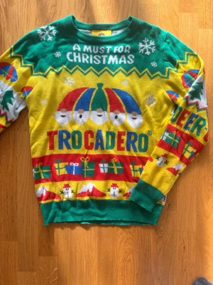 Färgglad jultröja Trocadero - Säljer en färgglad stickad jultröja från Trocadero med grönt, gult, rött och blått. Tröjan har motiv av tomtar, snögubbar, granar, presenter och texten 'A must for Christmas'. Perfekt för dig som vill sticka ut på julfesten!