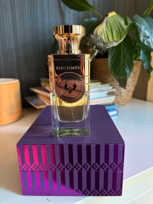 Electimuss Imoerium 100ml Extrait de Parfum  - Snygg parfym från Electimuss i en exklusiv glasflaska med guldfärgat lock och detaljer. Flaskan har en elegant, rektangulär form och dekoreras med ett stilrent emblem. Kommer i en lila ask med metalliskt mönster. Perfekt för dig som gillar lyxiga dofter och snygg design.