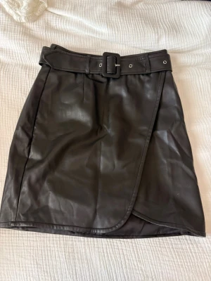 Svart omlottkjol i skinnimitation - Snygg svart kort omlottkjol i skinnimitation med bälte i midjan och markerat spänne. Köpt för flera år sen, använd max 2 gånger. 