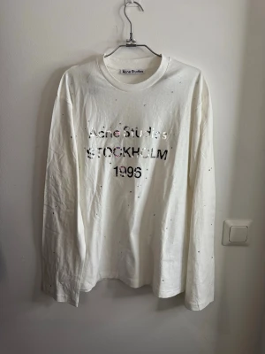 Vit långärmad t-shirt Acne Studios - Vit långärmad t-shirt från Acne Studios med tryck i silvertext 'STOCKHOLM 1996' på bröstet. T-shirten har små glittriga detaljer över hela framsidan och är gjord i mjuk bomull. Perfekt för dig som gillar stilrena plagg med en twist.