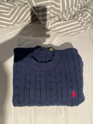 Mörkblå kabelstickad tröja Polo Ralph Lauren - Snygg mörkblå kabelstickad tröja från Polo Ralph Lauren med klassisk röd logga broderad på bröstet. Tröjan har rund halsringning, långa ärmar och ribbade muddar. Perfekt för dig som gillar tidlös och stilren design.