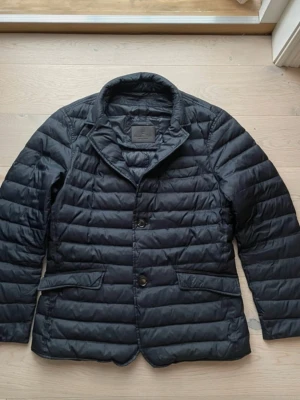 Massimo Dutti jacka - Stilren svart dunjacka med quiltad design och två knappar framtill. Jackan har klassisk krage och två fickor med lock. Perfekt för dig som vill ha en lätt och snygg jacka till vardags. M/L