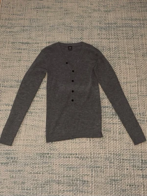 Grå kofta med knappar från MQ - Stilren grå cardigan från MQ med v-ringning och svarta knappar framtill. Tillverkad i mjukt, ribbstickat material och har långa ärmar. Perfekt att slänga över en t-shirt för en enkel och snygg look.