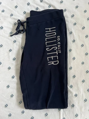 Mörkblå vintage low rise mjukisbyxor Hollister - Snygga vintage low rise låg midja mörkblå mjukisbyxor från Hollister med stort vitt tryck längs benet där det står 'HOLLISTER SO CALIF'. Byxorna har dragsko i midjan och muddar vid bensluten. Perfekta för chill dagar och riktigt sköna att ha på sig. Storlek M men stretchiga och snöre så passar xs-m. Måtten finns. 