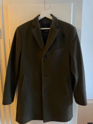 Mörkgrön rock från J.Lindeberg - Stilren mörkgrön rock från J.Lindeberg med klassisk krage och tre knappar framtill. Rocken har två snedställda fickor och en bröstficka. Tillverkad i ett tjockare tyg som passar perfekt för kyligare dagar.