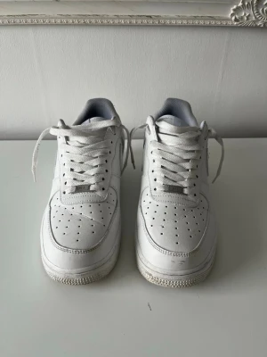Vita Nike Air Force 1 sneakers - Klassiska vita Nike Air Force 1 sneakers i lågt utförande. Skorna har perforerad tå för ventilation, vita skosnören och en robust sula. Tillverkade i läder med textildetaljer på insidan. Perfekta för en clean och stilren look.