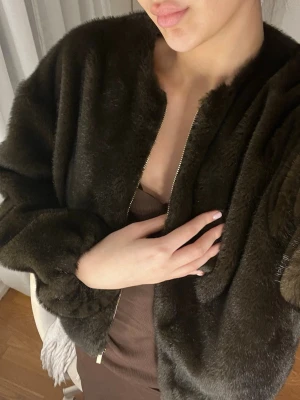 Mörkbrun fuskpälsjacka med dragkedja - Snygg mörkbrun fuskpälsjacka med dragkedja framtill och rundad hals. Jackan har en mjuk och fluffig yta samt ballongärmar för en trendig look. Perfekt för att hålla värmen under kalla dagar och ger en lyxig känsla till outfiten.