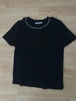 Svart t-shirt med pärldetaljer - Stilren svart t-shirt från Zara med korta ärmar och rund halsringning dekorerad med vita pärlor. T-shirten har en enkel och klassisk passform, perfekt för dig som gillar minimalistiska detaljer. Materialet känns mjukt och bekvämt.