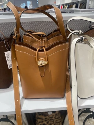 Brun bucket bag med gulddetalj - Snygg brun axelväska i bucket-modell med justerbar axelrem och guldfärgad vridknapp framtill. Väskan har en stilren design med vita sömmar och är gjord i slätt konstläder. Perfekt för dig som gillar trendiga accessoarer med en twist.