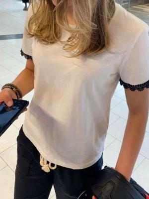 Vit t-shirt med svart spetskant - Snygg vit t-shirt i bomull med korta ärmar och svart spetsdetalj vid ärmsluten. Enkel och stilren design som passar perfekt till jeans eller shorts. T-shirten har en normal passform och känns både fräsch och trendig.