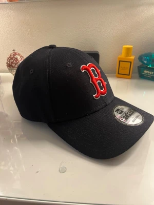 Svart New Era Boston Red Sox keps - Säljer en svart keps från New Era med Boston Red Sox-logga i rött och vitt framtill. Klassisk böjd skärm och broderad New Era-logga på sidan. Justerbar baktill och tillverkad i bomullstyg. Perfekt för dig som gillar sportig streetstyle.