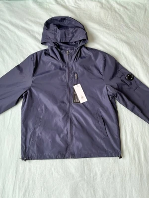 Mörkblå vindjacka från C.P. Company - Snygg mörkblå vindjacka från C.P. Company med huva, dragkedja framtill och två sidofickor. Jackan har en ikonisk linsdetalj på ärmen och är tillverkad i lätt, vattenavvisande material. Perfekt för blåsiga dagar och streetwear-stil.