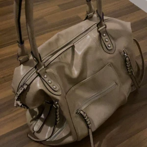 Beige handväska med flätade detaljer - Snygg beige handväska i mjukt konstläder med flätade detaljer på dragkedjorna och dubbla handtag. Väskan har en stor ytterficka med dragkedja och dekorativa nitar vid handtagsfästena. Perfekt för dig som gillar stilrena och praktiska accessoarer. Säljer för den passade inte till mina kläder❤️