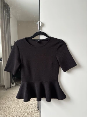 Svart peplumtopp med struktur - Snygg svart peplumtopp med rund halsringning och halvlånga ärmar. Toppen har en strukturerad yta och markerad volang nedtill som ger en feminin siluett. Perfekt att styla med jeans eller kjol för en trendig look.  ‼️Kan visa bild hur den ser ut på & Pris kan diskuteras!!