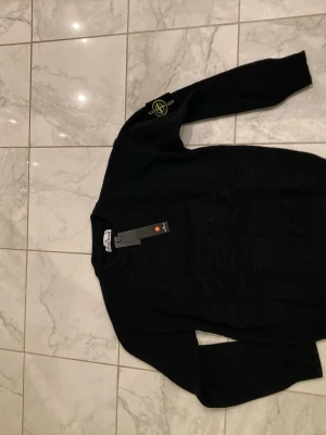 Svart stickad tröja från Stone Island - Snygg svart stickad tröja från Stone Island med den klassiska loggan på vänster ärm. Tröjan har rund halsringning och ribbade muddar vid ärmslut och nederkant. Perfekt för dig som gillar streetwear och vill ha något stilrent och bekvämt.