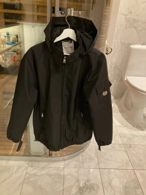 Svart vindjacka från Moncler - Snygg svart vindjacka från Moncler med huva och dragkedja framtill. Jackan har två fickor med dragkedja och en ficka med knapp på ärmen där Moncler-loggan sitter. Tillverkad i ett lätt och tåligt material som passar perfekt till blåsiga dagar.