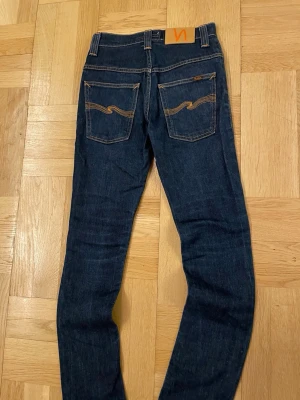Mörkblå jeans från Nudie Jeans - Säljer ett par mörkblå jeans från Nudie Jeans med slim passform och är herr. Jeansen har kontrastsömmar i orange och Nudie-logga på bakfickan samt läderpatch i midjan. Tillverkade i slitstark denim av bomull. Perfekta för dig som gillar stilrena och tidlösa jeans.
