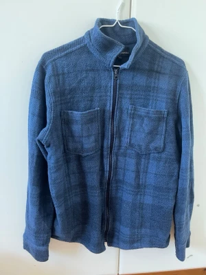 Blå overshirt med dragkedja - Snygg blå overshirt från KappAhl med diskret rutmönster. Modellen har två bröstfickor, hög krage och dragkedja framtill. Tillverkad i mjukt tyg som ger en avslappnad look. Perfekt att slänga över en t-shirt för en cool lager-på-lager-stil.