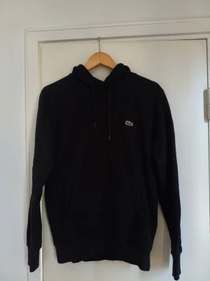 Svart hoodie från Lacoste - Säljer en svart hoodie från Lacoste med klassisk krokodillogga på bröstet. Hoodien har dragsko i huvan och en stor magficka. Tillverkad i mjuk bomullsblandning och har ribbade muddar vid ärmslut och nederkant. Perfekt för en avslappnad och stilren look.Hoodien är som ny i storlek Medium.