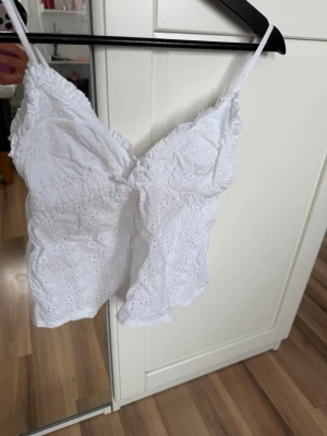 Vit broderad linnetopp med spets - Söt vit linnetopp med tunna axelband och broderade detaljer. Toppen har v-ringning, spets och justerbara axelband. Perfekt för varma dagar och ger en fräsch sommarkänsla.