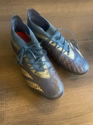 Adidas Predator blå fotbollsskor - Snygga Adidas Predator fotbollsskor i blått med silvriga och röda detaljer. Skorna har snörning, högre strumpkrage och mönstrad ovandel för bättre bollkontroll. Perfekta för dig som vill sticka ut på planen och få grym touch på bollen. Nypris 2799 obs ens skons underläggs färg har försvunnit men sulan e kvar! Inget som spelar roll.