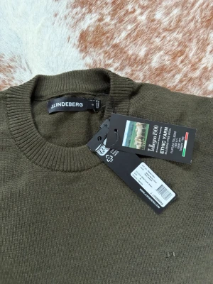 Grön Merino från J.Lindeberg - Snygg olivgrön stickad tröja från J.Lindeberg i tunn merinoull. Tröjan har rund halsringning, ribbade muddar och diskret logga broderad framtill. Perfekt för dig som gillar stilrena och klassiska plagg med en modern touch. Nypris 1400. Helt ny, då jag  köpte fel storlek. 100% Merino. 