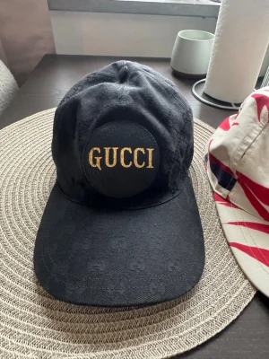 Svart keps från Gucci med logga - Snygg svart keps från Gucci med stor rund patch framtill där det står GUCCI i guldiga bokstäver. Klassisk böjd skärm och justerbar baksida. Materialet ser ut att vara bomull med diskret mönster i tyget. Perfekt för dig som vill ha en stilren och lyxig accessoar.