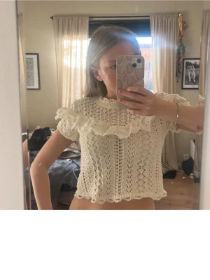 Krämvit stickad blus med volang - Supersöt vit stickad blus från Zara med korta ärmar och volangdetalj över bröstet. Blusen är croppad och har ett fint spetsmönster som ger en romantisk vibe. Perfekt att matcha med jeans eller kjol för en trendig look.