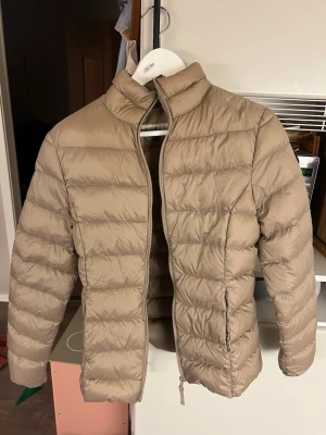 Beige dunjacka Hangarun Republic - Snygg beige dunjacka från Hangarun Republic med patch på ärmen där det står N.Y USA och 27. Jackan har hög krage, quiltad design och dragkedja framtill. Perfekt för kalla vinterdagar och riktigt lätt att bära.