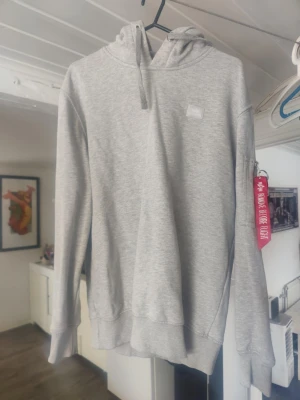 Ljusgrå hoodie Alpha Industries - Snygg ljusgrå hoodie från Alpha Industries med klassisk huva och dragsko. Har en ficka med dragkedja och röd tag på ena ärmen. Tillverkad i mjukt sweatshirtmaterial, perfekt för en avslappnad streetwear-look. Ena snör brickan har lossat 