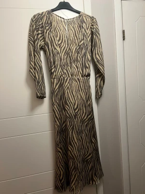 Adoore Zebramönstrad långklänning - Adoore Cool långklänning med zebramönster i beige och brunt. Klänningen har långa ärmar och en smal passform upptill med lätt utsvängd kjol. Materialet känns mjukt och följsamt, perfekt för dig som vill sticka ut.
