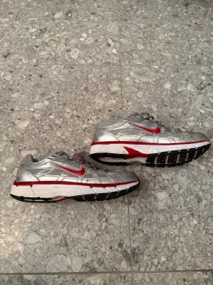Nike silver och röda sneakers - Säljer ett par Nike sneakers i silver med röda detaljer och vit sula. Skorna har mesh och syntetmaterial på ovandelen, klassisk Nike-logga på sidan och snörning. Perfekta för dig som gillar sportig stil och vill sticka ut med coola färger.