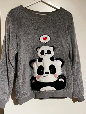 Grå loungewear-tröja med panda - Mysig grå loungewear-tröja med ett stort broderat pandamotiv på framsidan och en liten panda ovanpå med ett hjärta. Tröjan är gjord i mjukt fleecematerial och har rund hals och långa ärmar. Perfekt för hemmamys och chill.