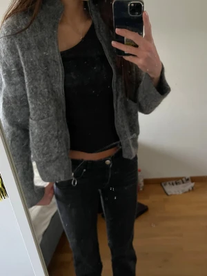 Grå zip-kofta med fickor - Mysig grå kofta med dragkedja och rund ringdetalj i metall. Koftan har två stora fickor framtill och är tillverkad i ett fluffigt, stickat material. 