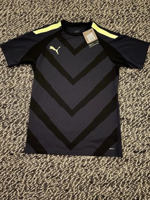 Svart sporttröja från Puma - Snygg svart sporttröja från Puma med korta ärmar och gul logga på bröstet. Tröjan har ett coolt V-mönster framtill och gula detaljer på axlarna. Tillverkad i ett lätt och snabbtorkande material, perfekt för träning och aktivitet.