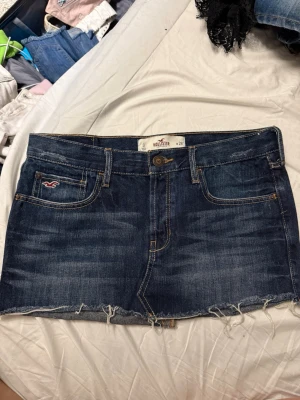 Mörkblå jeanskjol från Hollister - Säljer en snygg mörkblå jeanskjol från Hollister med rå kant nertill och klassiska fem fickor. Kjolen har en normal passform och är perfekt för dig som gillar en avslappnad och trendig stil. Ikonisk Hollister-logga på fickan framtill.