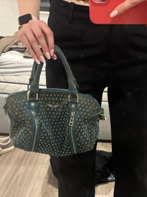 Blå Zadig&Voltaire handväska med nitar - Cool blå handväska från Zadig&Voltaire i mocka med silvriga metalldetaljer och små nitar. Väskan har dubbla handtag, två dragkedjor framtill och en rymlig insida. Perfekt för dig som gillar edgy och trendiga accessoarer.