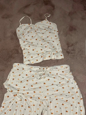 Vit pyjamas med apelsinmönster lager 157 - !!!!!!!Byxor är i M, toppen är i XS Söt pyjamas med vitt linne och matchande byxor, båda med små apelsinmönster. Linnet har tunna axelband och byxorna har resår och dragsko i midjan för extra komfort. Perfekt för mysiga kvällar hemma. !!!!!!!!!!Byxor är i M 