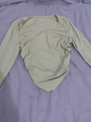 Beige ribbad långärmad topp - Trendig beige långärmad topp med ribbad struktur och bred, rundad urringning. Toppen har en figurnära passform och lätt rynkade sidor som ger en snygg siluett. Perfekt att styla med jeans eller kjol för en cool vardagslook.