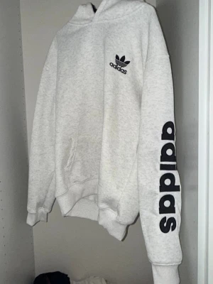 Grå hoodie från Adidas Originals - Snygg ljusgrå hoodie från Adidas Originals med stor svart logga på huvan och mindre logga på bröstet. Ärmen har Adidas-tryck i svart. Klassisk känguruficka framtill och ribbade muddar. Tillverkad i mjuk bomullsblandning, perfekt för en avslappnad streetwear-look.