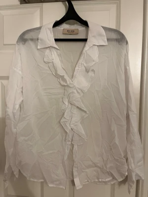 Vit volangblus från Mos Mosh - Snygg vit blus från Mos Mosh med volangdetaljer längs knäppningen och klassisk krage. Blusen har lång ärm och är tillverkad i ett tunt, lätt material som ger en luftig känsla. Perfekt att styla med jeans eller kjol för en trendig look.