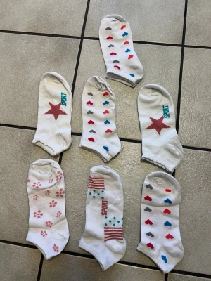 7 par Vita låga strumpor med mönster storlek 36-38 - Säljer ett gäng vita låga strumpor med olika mönster som hjärtan, stjärnor, tassavtryck och ränder. Några har texten 'SPORT' i färg. Färgerna på mönstren är röd, blå, grå och rosa. Perfekta till sneakers eller låga skor.