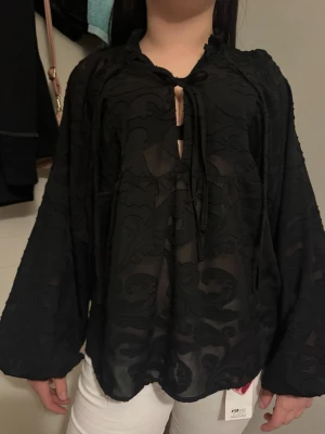 Svart mönstrad blus med knyt - Snygg svart blus med transparent mönster och vida långa ärmar. Blusen har knytning framtill vid halsen och ett lätt genomskinligt tyg som ger en cool look. Perfekt att styla med jeans eller kjol för en trendig outfit.