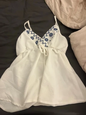 Vit broderad linnetopp med knyt - Söt vit linnetopp med tunna axelband och blå broderade blommor vid urringningen. Toppen har v-ringad hals, knyt i midjan och är tillverkad i ett lätt, luftigt material. Perfekt för varma dagar och enkel att styla med jeans eller kjol.