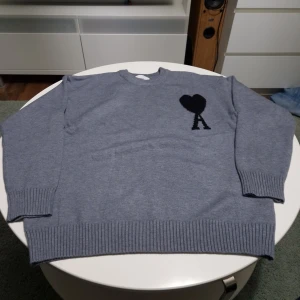 Grå stickad tröja Ami Paris - Säljer en grå stickad tröja från Ami Paris med svart hjärta och bokstaven A broderat på bröstet. Tröjan har rund hals, ribbade muddar och är tillverkad i mjukt material. Perfekt för dig som gillar stilrena och trendiga plagg.