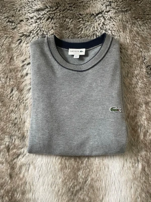 Grå tröja från Lacoste S - Klassisk grå tröja från Lacoste med rund halsringning och den ikoniska gröna krokodilloggan broderad på bröstet. Tröjan har ribbade muddar och är tillverkad i mjukt material som känns skönt mot huden. Perfekt för en clean och stilren look.