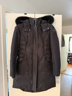 Svart dunjacka med pälshuva från Moose Knuckles - Snygg svart dunjacka från Moose Knuckles med avtagbar huva och svart äkta päls . Jackan har långa ärmar, dragkedja och tryckknappar framtill samt två stora fickor. Ikonisk logga på ärmen och quiltade partier på sidorna för extra stil.