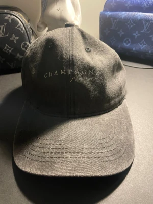 Svart keps Champagne Please - Säljer en svart keps med texten 'CHAMPAGNE please' broderad framtill. Klassisk böjd skärm och justerbar rem bak. Tillverkad i bomullstyg för en avslappnad och stilren look.