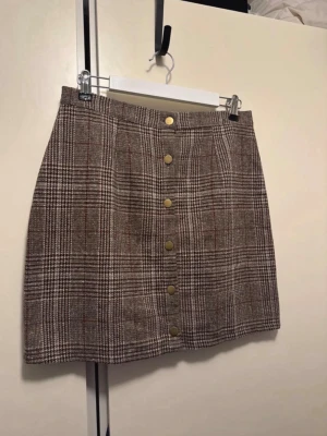 Rutig kortkjol med knappar framtill - Snygg rutig kortkjol i bruna och beige toner med dekorativa guldknappar framtill. Kjolen har en klassisk A-linjeform och är tillverkad i ett tjockare tyg som ger en stilren look. Perfekt att matcha med stickad tröja eller boots. Ingen storlekslapp men skulle gissa på att det motsvarar ca 42 då den funkade på mig.