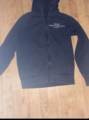 Svart hoodie Peak Performance zip - Svart hoodie från Peak Performance med dragkedja framtill och fickor på magen. Klassisk logga tryckt på bröstet. Mjuk bomullskänsla och huva med snörning. Perfekt för en avslappnad och sportig stil.